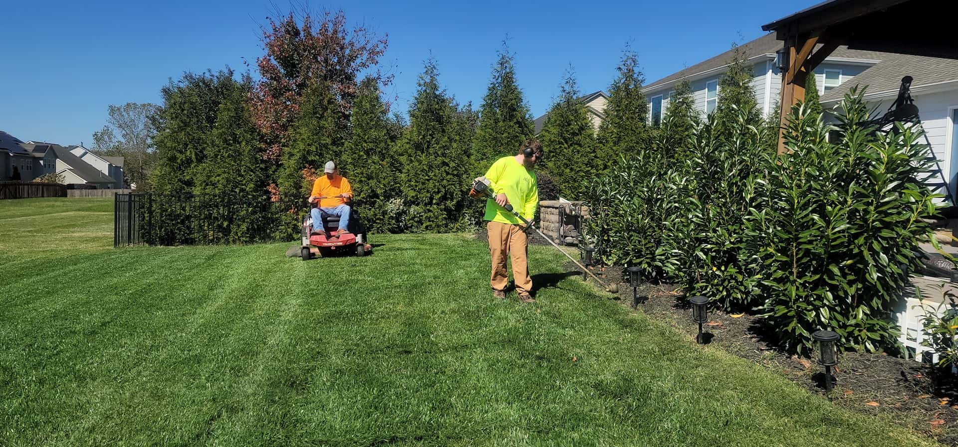 Crew member on a riding mower cutting fresh stripes into a green residential lawn, second worker trimming
